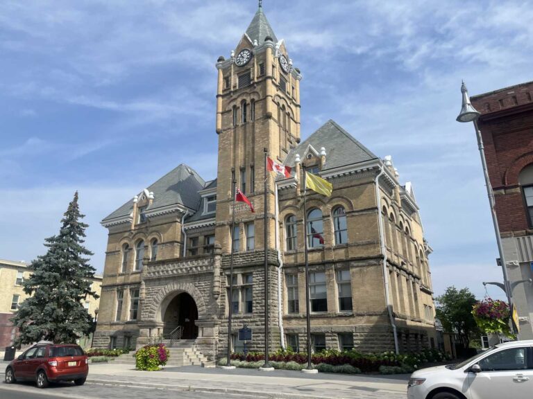 St. Thomas City Hall, a yellow-brick Victorian municipal building with clock tower, seen at an angle from Talbot Street with flags flying