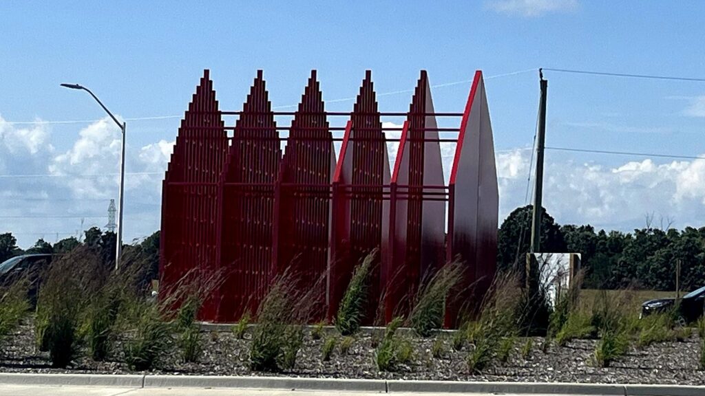 Fading Memories, the bright red barn-silhouette sculpture of vertical slats in the Highbury Avenue and Ron McNeil Line roundabout in St. Thomas, with ornamental grasses at its base