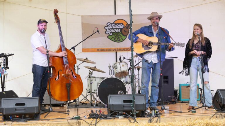 A trio on the hay-bale-fronted tent stage at the 2025 Middle River Music Fest at Common Ground Farm near St. Thomas: an upright bass, an acoustic guitar and a woman at the right-hand mic, beneath the festival banner.