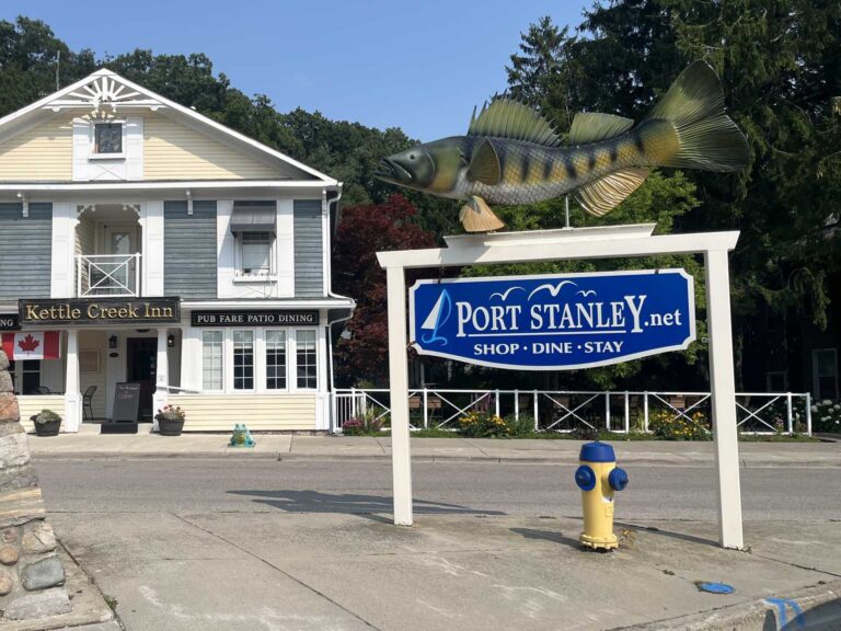 A large fish sculpture perches on top of the blue PortStanley.net Shop, Dine, Stay sign in the village core, with the historic Kettle Creek Inn behind.