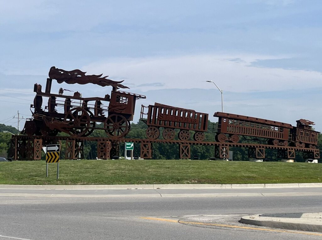 Rusted steel silhouette sculpture of a locomotive and rail cars lettered St. Thomas on a trestle base in the centre of a roundabout