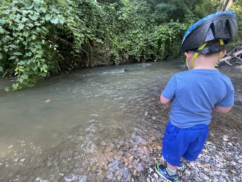 A young child wearing a bike helmet stands on the stony bank of Catfish Creek at the Yarmouth Natural Heritage Area near Sparta.