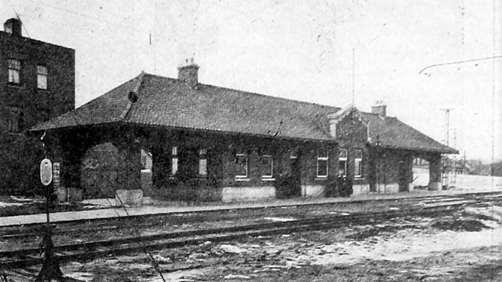 Black-and-white photograph, published in 1920, of a long, low brick railway station with a hipped roof and an open shelter at each end, seen across railway tracks.