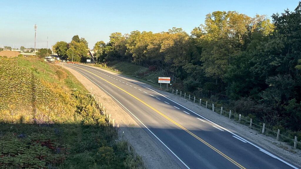 An empty two-lane Highway 3 curving east toward a line of orange construction barrels across the road near the First Avenue interchange, with a roadside sign reading Highway 3 Eastbound Closed, First Ave to Centennial, evening light