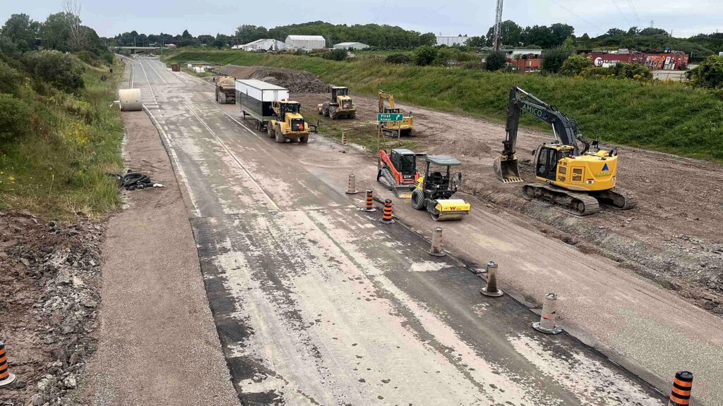 The closed stretch of Highway 3 in St. Thomas seen from the Burwell Road overpass looking west toward First Avenue: the roadway stripped to gravel, an excavator, a roller, a loader towing a trailer, a dump truck, a large concrete pipe section, orange barrels and a green First Avenue exit sign