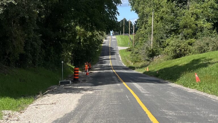Zoomed view along a rural paved road with a freshly gravelled shoulder and new asphalt edge on the left, striped construction barrels and cones beside a roadside drain grate, road open, yellow centre line rising to a crest with hydro poles