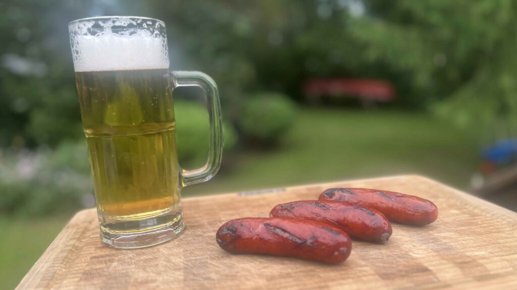A tall glass mug of lager with a thick head of foam beside three grilled sausages on a wooden cutting board, outdoors with a blurred green backyard behind