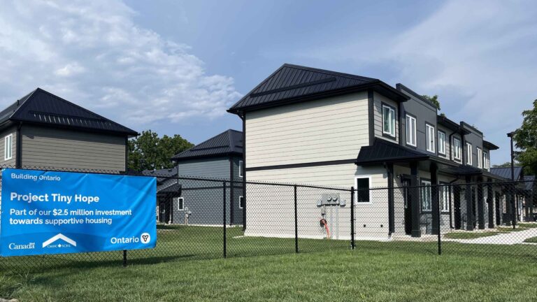 Two-storey housing units at Project Tiny Hope in St. Thomas behind a black chain-link fence, with a blue Building Ontario banner reading Project Tiny Hope, part of our 2.5 million dollar investment towards supportive housing.