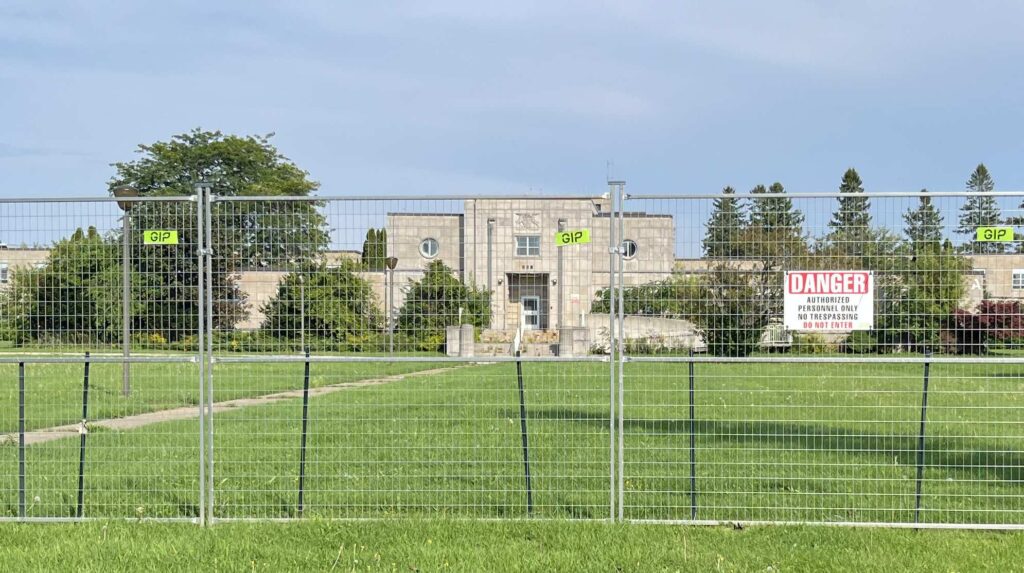 Temporary demolition fencing with a red DANGER Authorized Personnel Only sign in front of the limestone Art Deco administration building of the former St. Thomas Psychiatric Hospital.