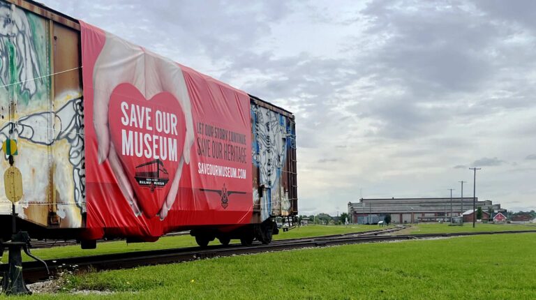 A red Save Our Museum banner showing a heart held in two hands covers the side of a graffiti-marked boxcar on a rail line in St. Thomas; the former Michigan Central locomotive shops that house the Elgin County Railway Museum stand in the distance under a cloudy sky