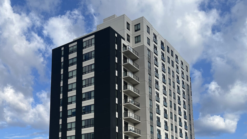 The upper floors of a 10-storey-plus apartment tower in St. Thomas, black and light-grey cladding with stacked balconies, against a blue sky with white clouds.