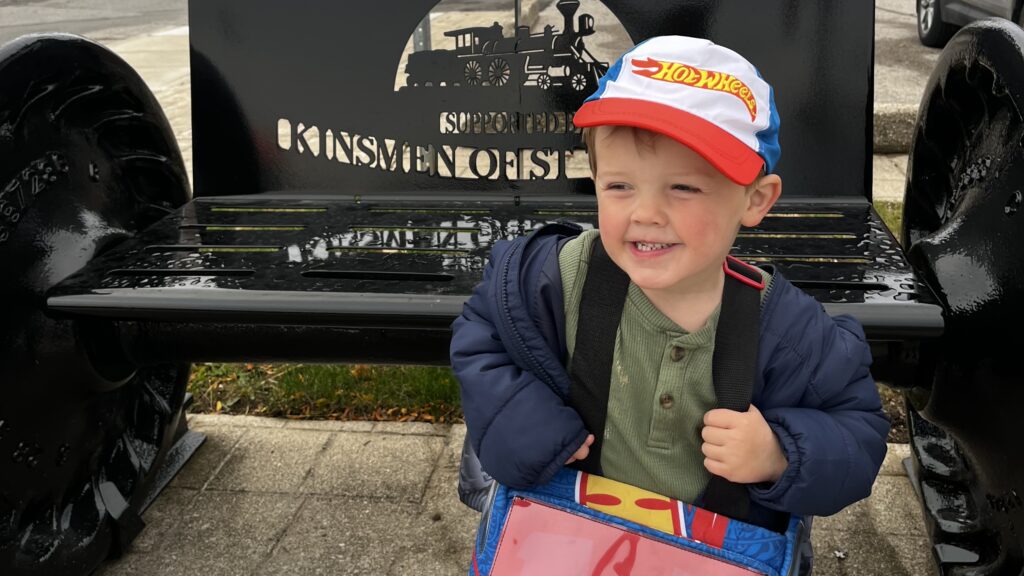 A smiling young child in a Hot Wheels cap and a toy race-car costume stands in front of a black railway-wheel bench in downtown St. Thomas at the 2024 Downtown Pumpkin Prowl.