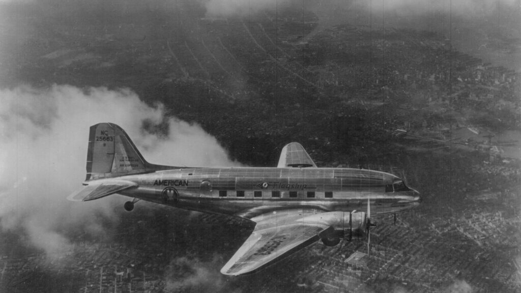 Black-and-white aerial photo of a silver twin-engine American Airlines DC-3 airliner, tail number NC 25663, flying above a city with clouds behind it