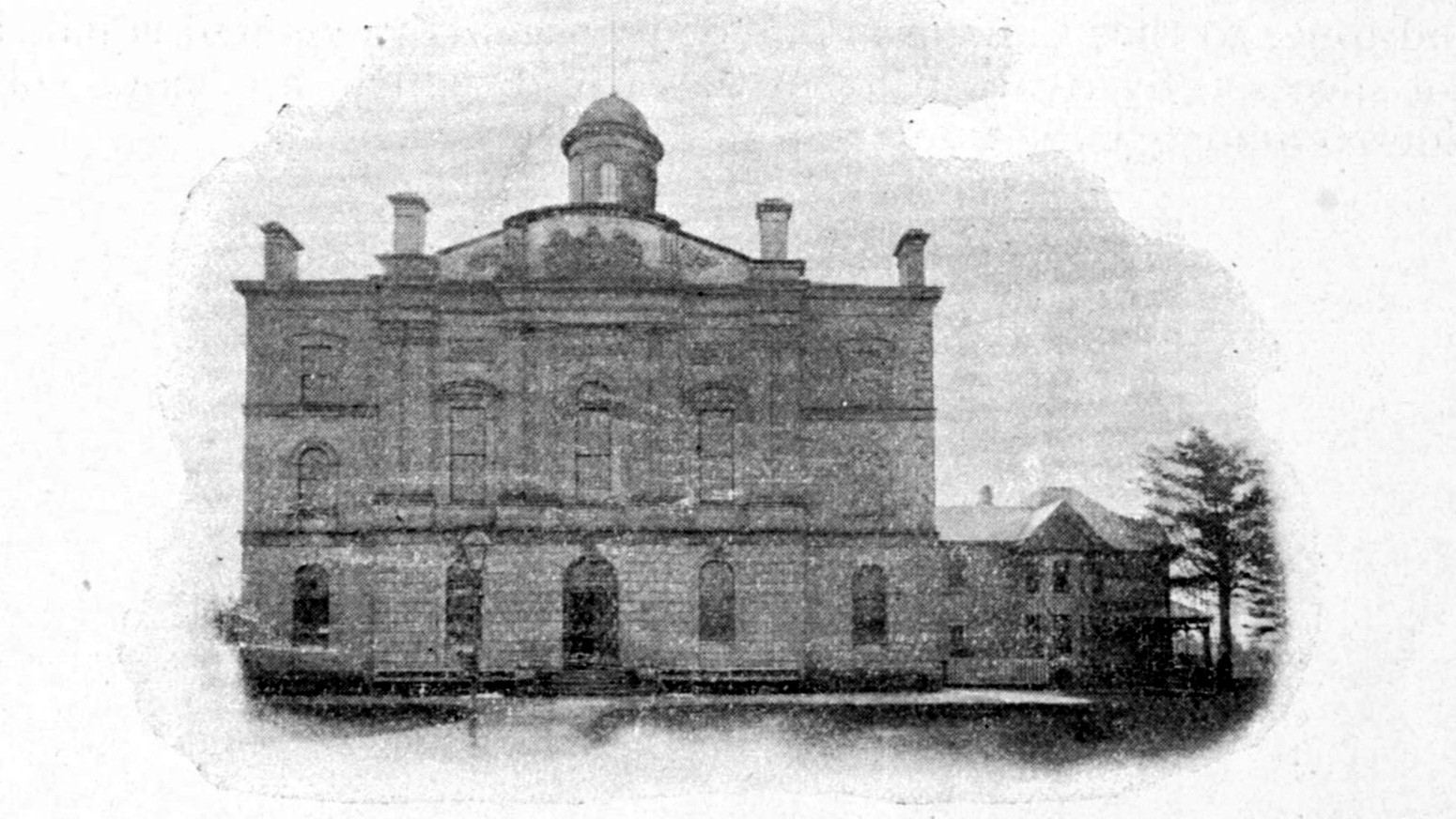 Black-and-white photograph, printed in 1901, of a three-storey stone courthouse with a small domed cupola and four chimneys, seen from the front, with a lower building and an evergreen tree to its right.