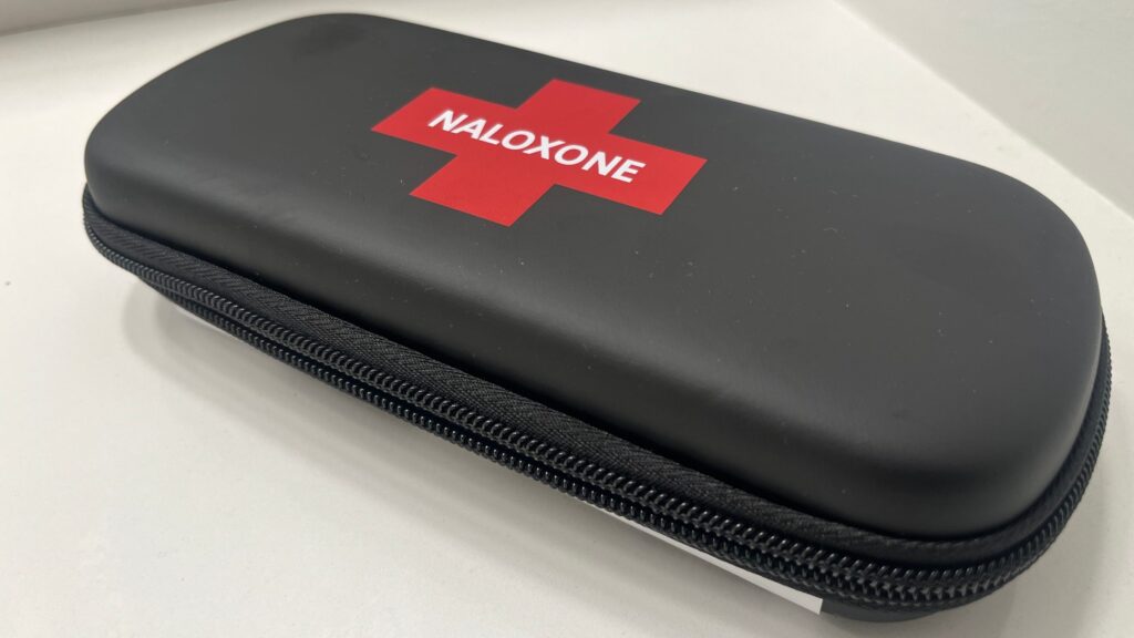 A closed black zippered naloxone kit case with a red cross reading NALOXONE, on a white counter