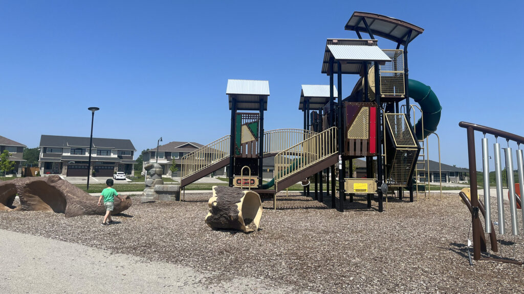 A playground with a tall climbing structure, log tunnel, rock sculpture and chimes on a wood-chip surface, a child walking at left, and new two-storey houses across the street behind it, in St. Thomas