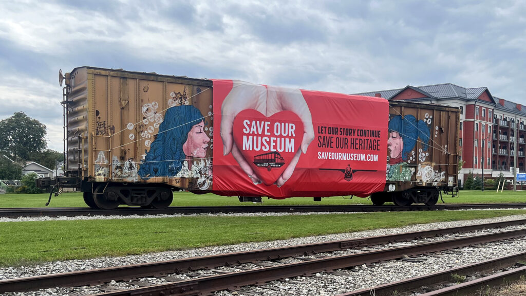 A red Save Our Museum banner with the Elgin County Railway Museum logo strapped to the side of a rust-brown boxcar painted with portraits of women with blue hair, on a rail siding in St. Thomas, September 2026