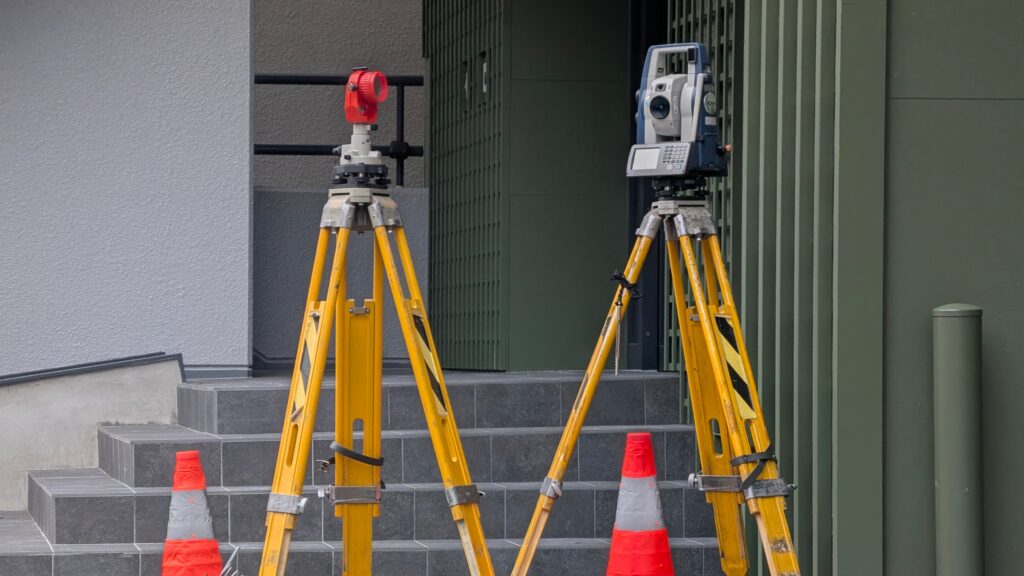 Two yellow surveying tripods, one holding a red survey prism and one a total station, beside orange traffic cones in front of the steps of a building