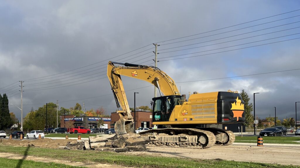 An excavator stands on the dug-up south side of Talbot Street near First Avenue in St. Thomas, with traffic on the north side of the road and businesses behind.